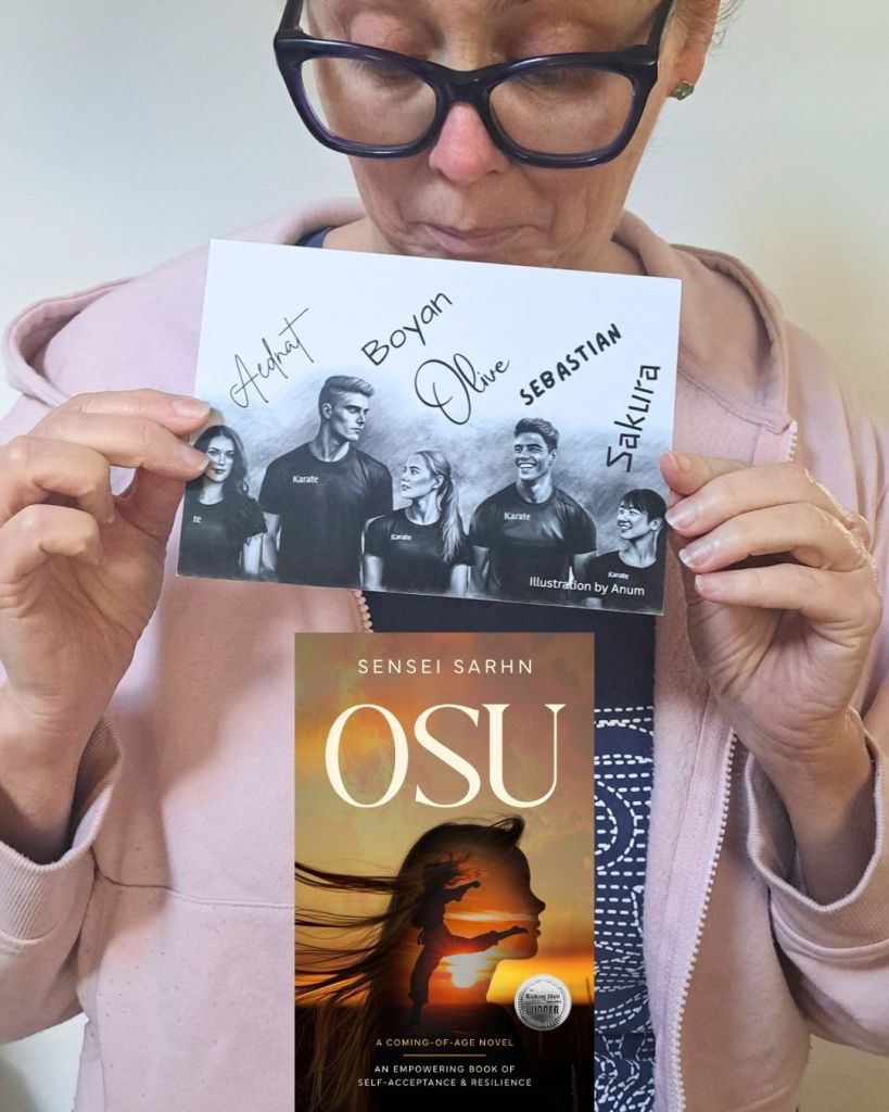 Australian author Sensei Sarhn, holds up a card with a drawing of the main characters of her empowering YA teen fiction novel, 'OSU'