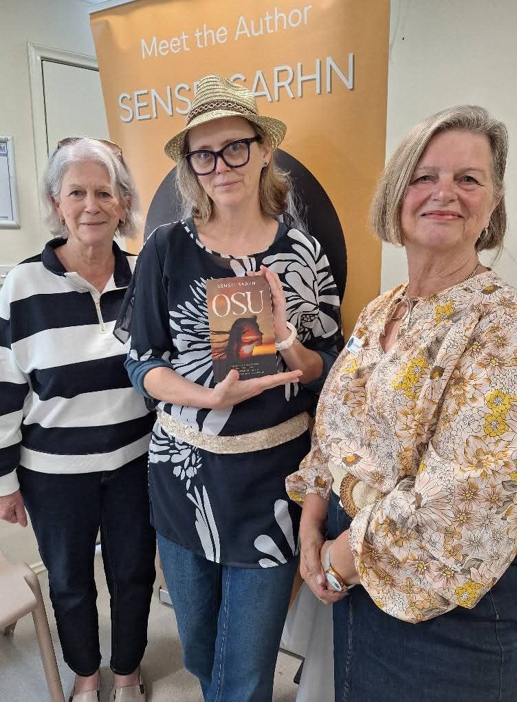 Australian female author, Sensei Sarhn wearing a hat stands in front of her banner, holding her young adult YA teen fiction and empowering coming-of-age novel 'OSU' beside two ladies from the CWA Picton District Branch